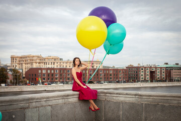 An attractive girl in a beautiful red dress sits on a pier by the river and holds huge coloured...