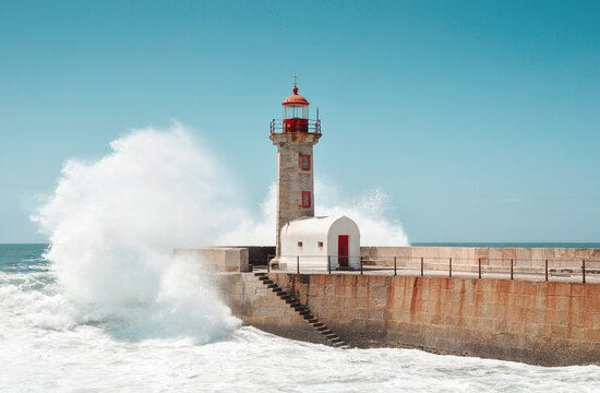 Wave crashing into lighthouse on the stone mole. The Felgueiras Lighthouse at Douro river mouth in Foz do Douro near Porto. Blue sky and Atlantic Ocean in the background.