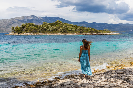 Young woman standing on a rocky beach is enjoying the beautiful view of the turquoise sea and a green island