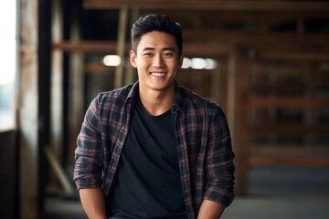 Fototapeta premium Portrait of a smiling asian man in his 20s wearing a comfy flannel shirt over empty modern loft background