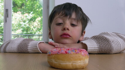 A young boy peeking over a table, eyes fixed on a colorful sprinkled donut, conveying the theme of...