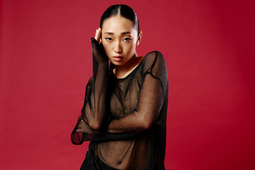 Asian woman in sheer top posing against red background with hands on hips and head tilted to the...