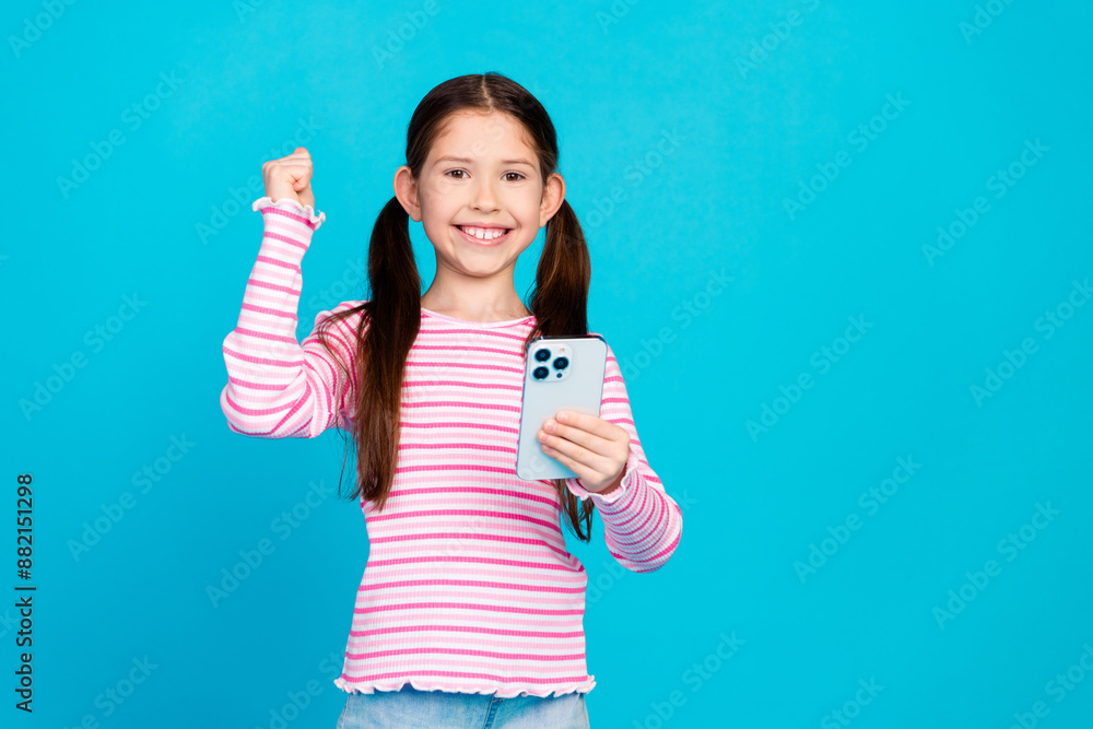 Poster Photo of cute girl with tails dressed striped shirt hold smartphone raising fist up win video game isolated on blue color background