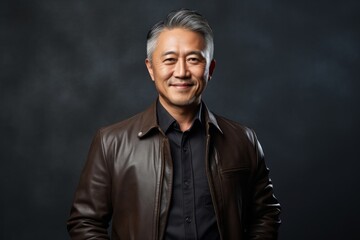Fototapeta premium Portrait of a grinning asian man in his 50s sporting a stylish leather blazer isolated on bare monochromatic room