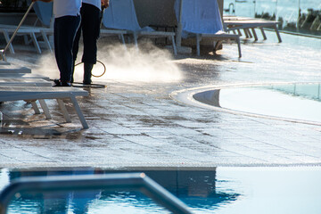 Worker Cleaning Poolside at Sunset, High-Pressure Water Spray Over Wet Floor, Sunlit Outdoor Swimming Pool