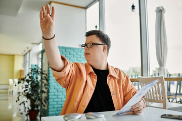 A man with Down syndrome sits at a table in a cafe, holding a menu and raising his hand to order.