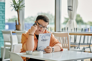 A man with Down Syndrome sits in a cafe, reviewing the menu while holding a phone to his ear.