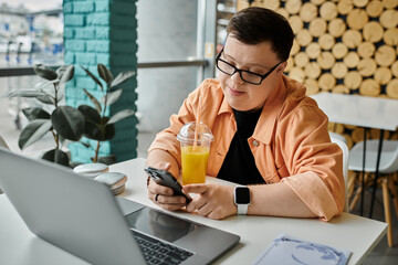 A man with Down syndrome sits at a cafe table, working on his laptop and holding a smartphone, while enjoying a beverage.