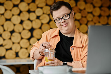 A man with Down syndrome smiles as he works on his laptop in a cafe, enjoying a refreshing drink during his workday.