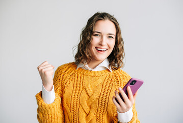 A joyful young woman celebrating good news on her smartphone. Standing against a white background,...