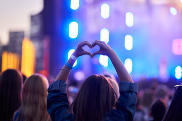 Back view of person forming heart shape with hands at music fest, surrounded by cheering crowd,...