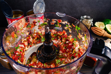 Freshly Made Blender Salsa in a Food Processor Bowl: Salsa in a food processor with empty tomato cans, spices, and a juiced lime in the background