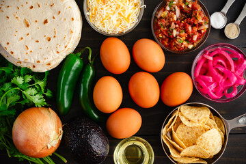 Tex-Mex Migas Breakfast Tacos Ingredients: Eggs, tortilla chips, jalapeno peppers, flour tortillas, shredded cheese, and more on a wooden table