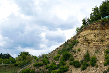 A hillside with a rocky, layered slope and green vegetation. The sky above is filled with white, puffy clouds.