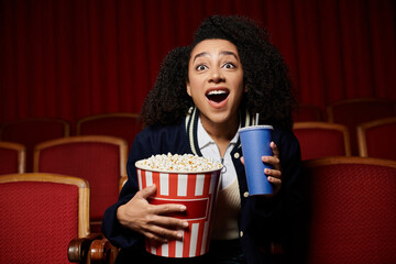 A young woman in a cinema watches a movie, holding a bucket of popcorn and a drink, reacting with excitement and surprise.