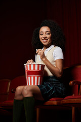 Naklejka premium A young woman enjoys a movie night, snacking on popcorn and reacting to the film with a big smile.