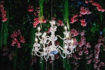 A luxury crystal chandelier decoration among artificial lush green foliage and pink flowers with blurred background