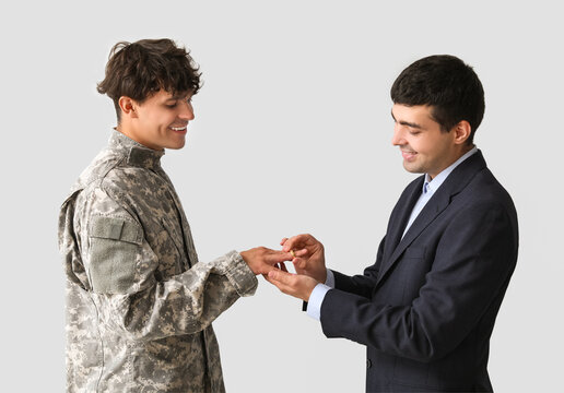 Young man putting wedding ring on his military husband's finger on light background