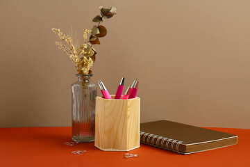 Wooden holder with pens, notebook and vase of dried flowers on red table