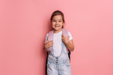 A girl with a backpack stands in front of a pink background.