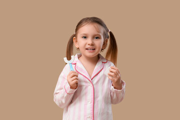 Happy little girl in pajamas with toothbrush and dental floss on brown background