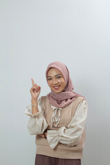 Portrait of young Asian woman in hijab in beige vest top with cheerful face with hands pointing up, white isolated