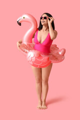 Beautiful young woman in stylish swimsuit with inflatable ring in shape of flamingo on pink background