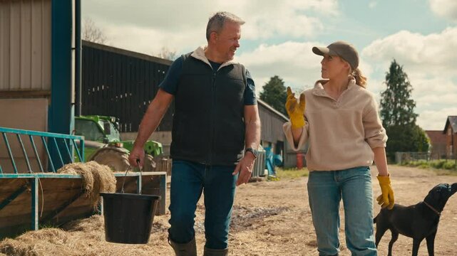 Male and female farm workers with dog walking across yard past cattle barn at feeding time carrying bucket  - shot in slow motion