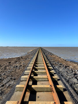 Straight railway tracks stretch along a stone dam or causeway into the sea toward the horizon under a clear blue sky.
