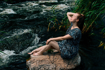 .Beautiful woman enjoying the river flow and fresh air while sitting on a stone in a blue dress