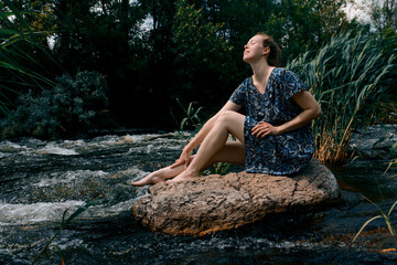 Beautiful woman in a blue dress strokes her legs in a river on a stone and smiles
