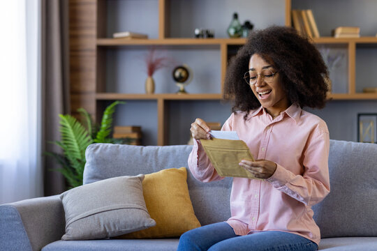 Joyful woman opening envelope while sitting on cozy sofa in comfortable home living room