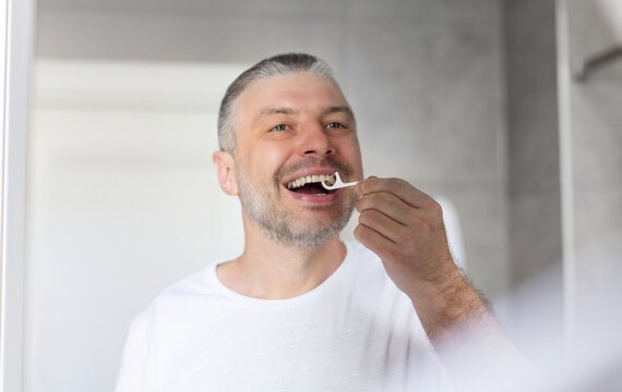 Middle aged man brushing his teeth using plastic toothpick with dental floss. Concept of hygiene and oral cavity