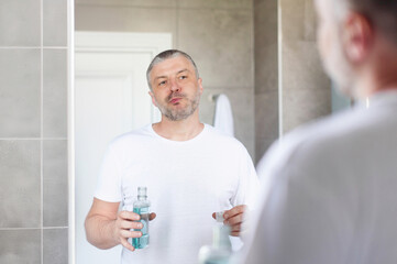 Middle aged man rinsing mouth with blue mouthwash, standing in bathroom in front of mirror. Teeth...