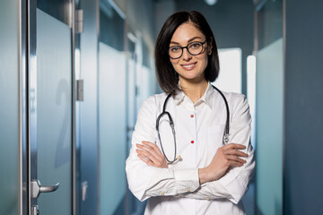 Smiling female doctor in white coat with stethoscope around neck standing with arms crossed in...
