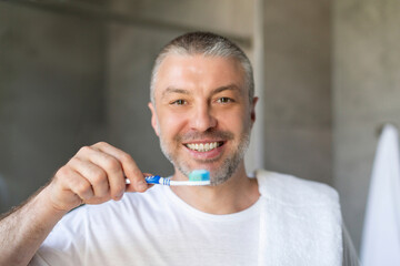Morning procedure and oral care. Middle aged man holding toothbrush with paste and smiling at camera in bathroom