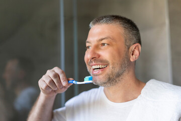 Positive European middle aged man with toothbrush in his hand smiling while brushing teeth in bathroom interior