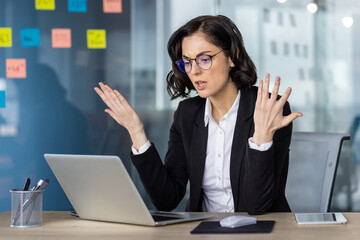 Angry woman in business attire expressing frustration while working on laptop in modern office...