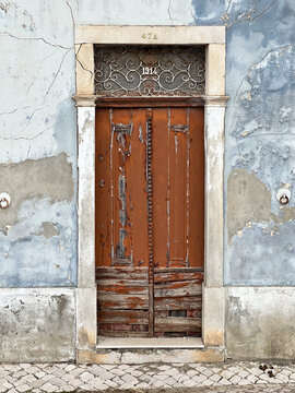 A weathered, rustic wooden door with peeling brown paint is framed by a stone lintel beneath a faded, cracking blue stucco wall along a cobblestone sidewalk in Azambuja Portugal.