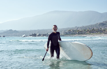 senior man comes out of the water holding a paddle board wearing surf suit