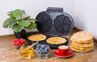 Making waffles at home. Waffle maker and stack of Belgian waffles with fresh berries, honey and cup of coffee on kitchen table. Cooking summer breakfast with heart-shaped waffles