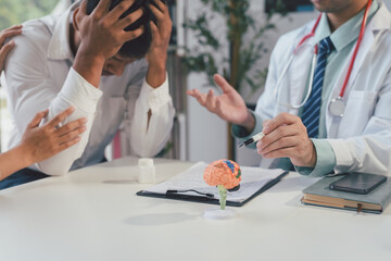 Doctor explaining something about a brain anatomical model to a male patient suffering from headache while a woman is comforting him