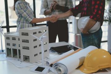 Team of architects is shaking hands in front of a model of a sustainable building with wind turbines and solar panels