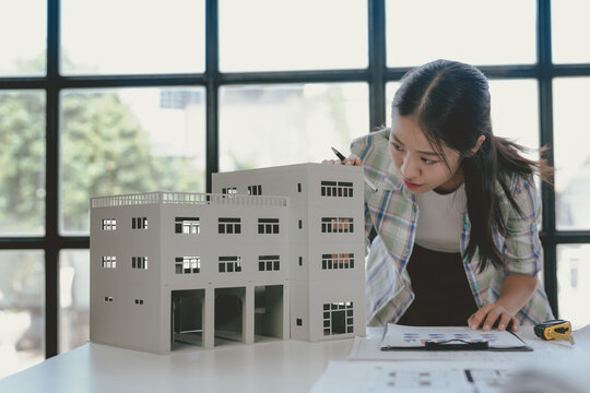 Young asian architect carefully reviews blueprints and uses a scale model to plan a new office building in the city