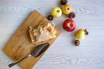 Piece of traditional thanksgiving apple cinnamon pie on the wooden cutting board with apples and walnuts on the white table. Homemade baked fruit tart with golden crust. Close up, copy space, top view