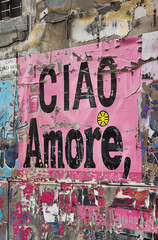 A pink sheet of paper with the words 'ciao amore' written in black letters is affixed to an old wall adorned with posters and stickers.