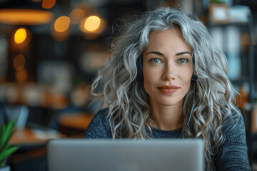 Smiling Middle Aged Woman with Gray Curly Hair Using Headset for Video Call with Customer at Desk in Modern Office