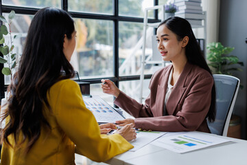 Two cheerful asian office workers women cooperating on project, sitting at desk with laptop using calculator, finance accounting analytical concept.