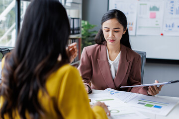 Obraz premium Two cheerful asian office workers women cooperating on project, sitting at desk with laptop using calculator, finance accounting analytical concept.