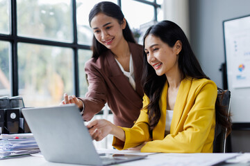 Two cheerful asian office workers women cooperating on project, sitting at desk with laptop using calculator, finance accounting analytical concept.